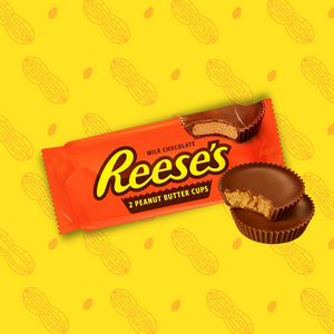 Sur cette image apparaît l'emblématique snack sucré salé américain. Les Reese's Cups sont conditionnées dans un emballage rectangulaire de couleur orange. Deux cups de Reese's sont placées devant l'emballage, ces gourmandises sont rondes et fourrées à la cacahuète.