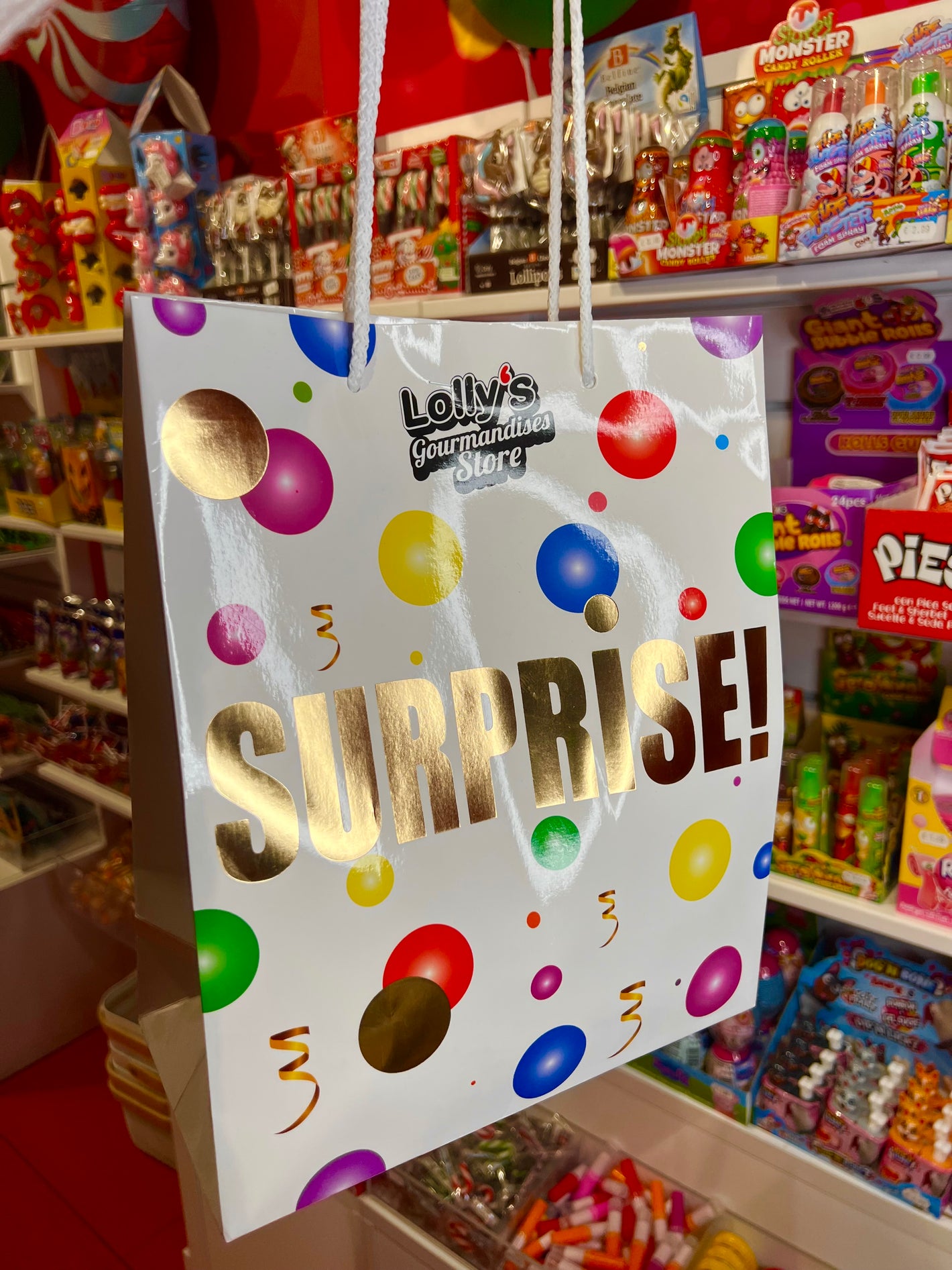 Sac surprise Lolly's, il possède des formes rondes colorées. L'incription "surprise" apparait au centre du sac de couleur or. 